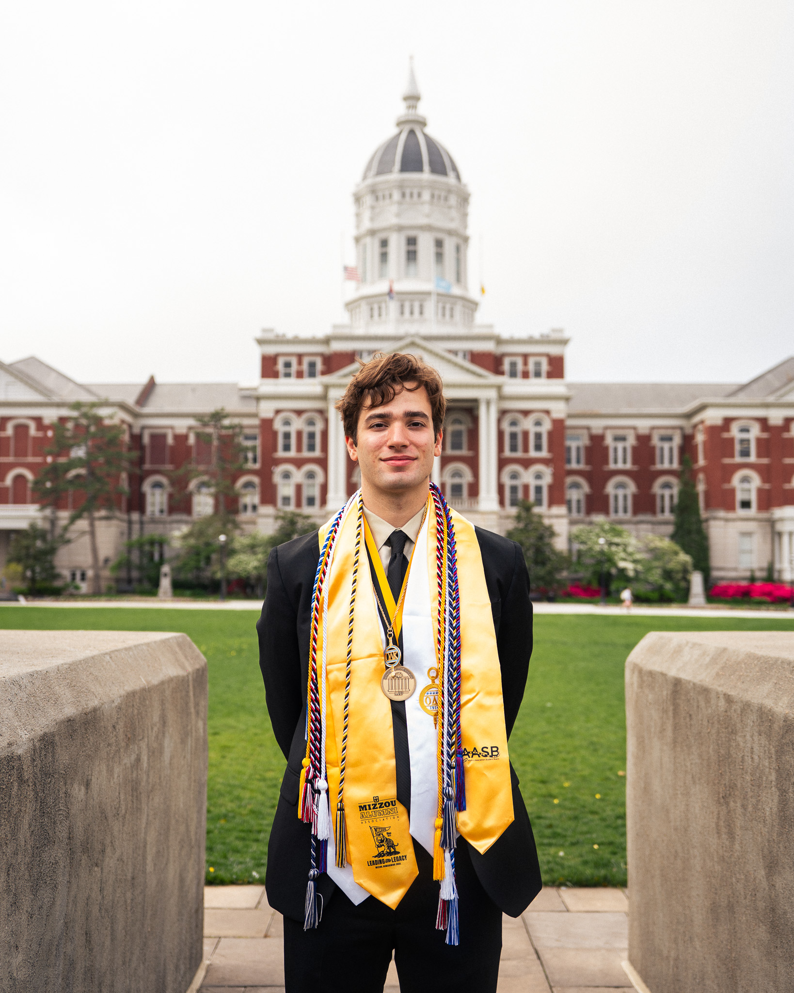 Logan Liguore in graduation stoles and cords stands in between the columns in front of Jesse Hall.